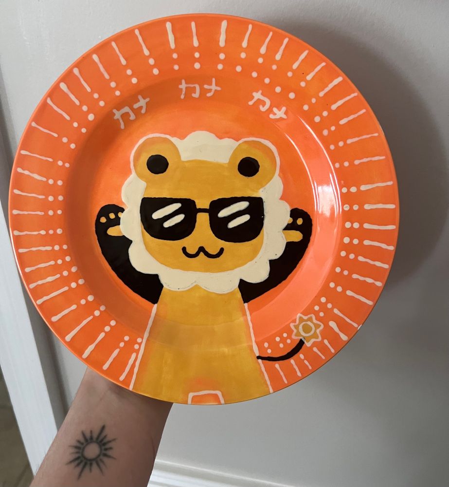 A small, orange plate painted with Rain’s mascot, Kana in the middle. The katakana for her name is repeated three times above her.