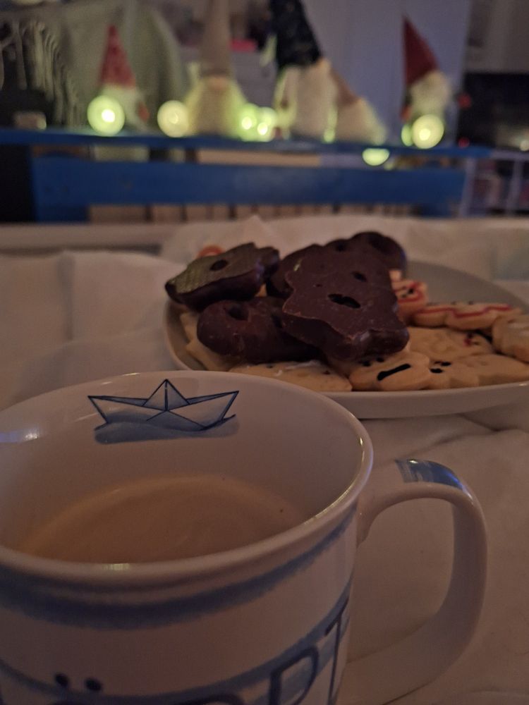 Eine blauweißgestreifte Kaffeetasse mit einem Papierschiff im Innenrand ist im Fokus. Dahinter sieht man einen Teller mit bunten Plätzchen und Lebkuchen. Im Hintergrund unschard ist ein blaues Regal zu sehen mit einer Lichterkette und 5 unterschiedlichen Wichteln.
