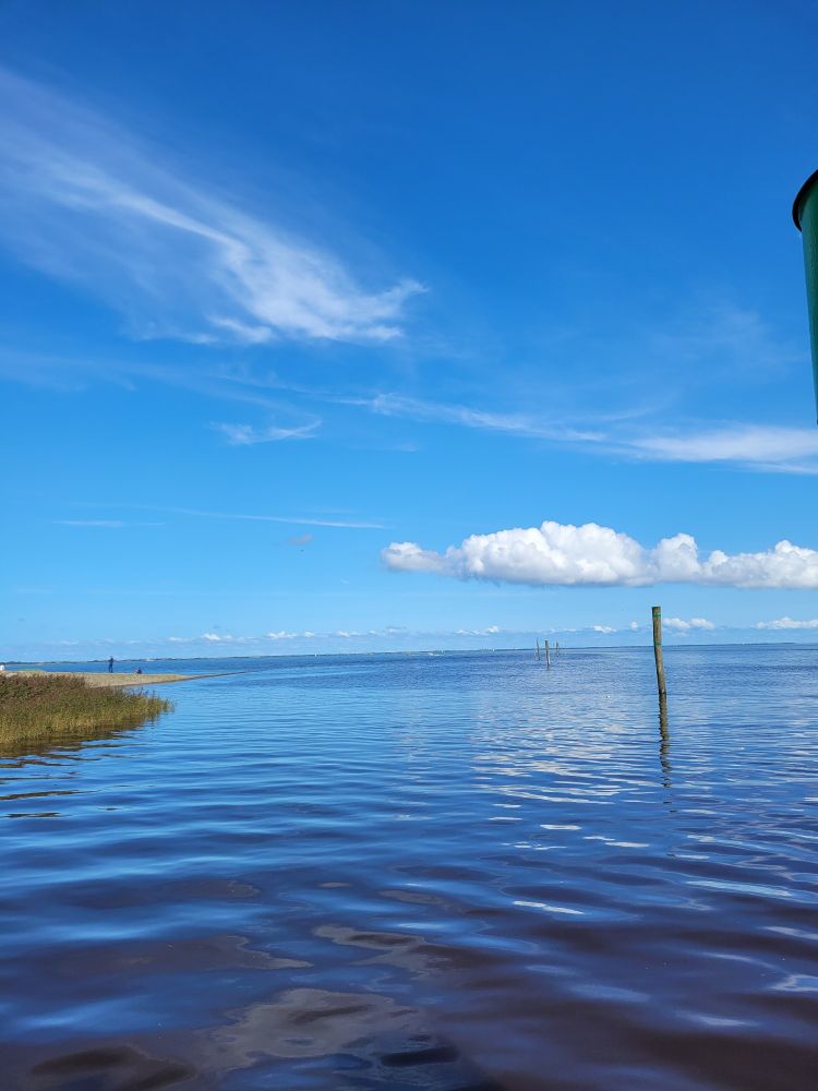 Man sieht das Meer. Rechts sind Holzpfähle zu sehen,  links ist eine mit Gras bewachsene Steinbuhne zu sehen. Der Himmel ist blau mit einzelnen Wolken am Horizont. Die Wolken spiegeln sich im Wasser.
