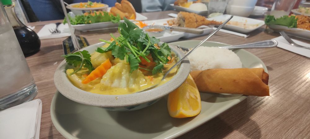Widescreen photo of a lunch set meal from a local Thai restaurant. There is a small bowl of yellow curry on top of a larger plate that holds a serving of white jasmine rice, an orange slice, and a fried egg roll. 