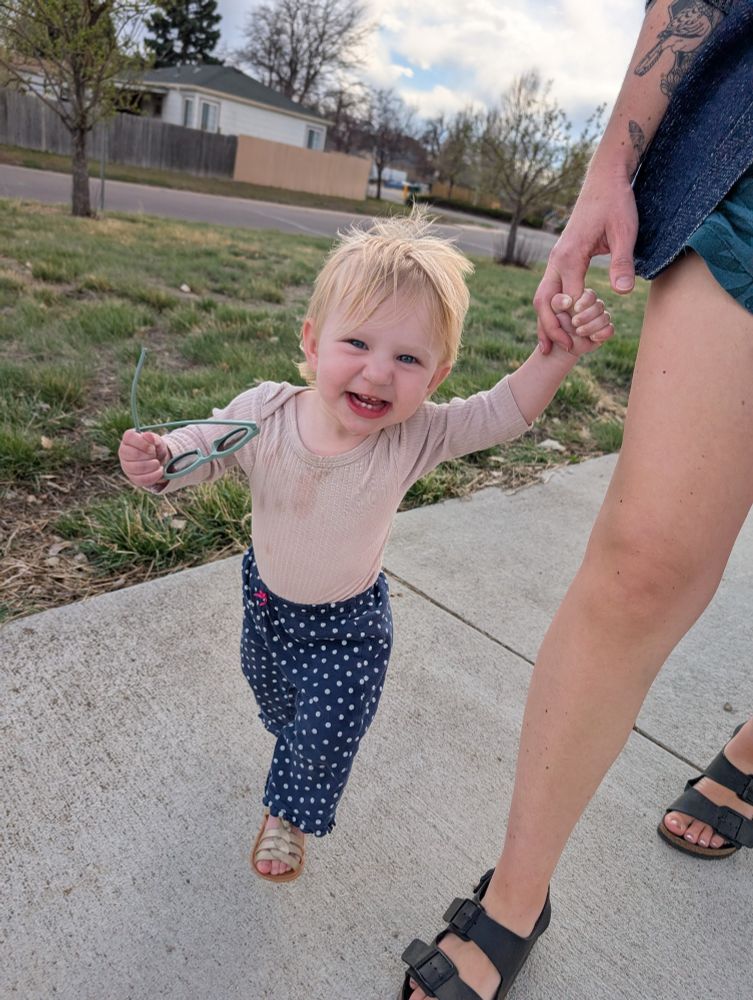 A baby walking holding her mom's hand in a particularly photogenic pose, smiling, with sunglasses in one hand 