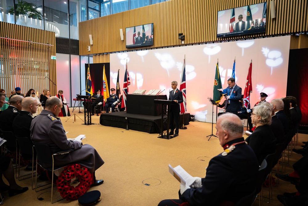 The Mayor of London speaking at the City Hall Remembrance Day service, with flags and poppy-themed lighting displayed behind him.