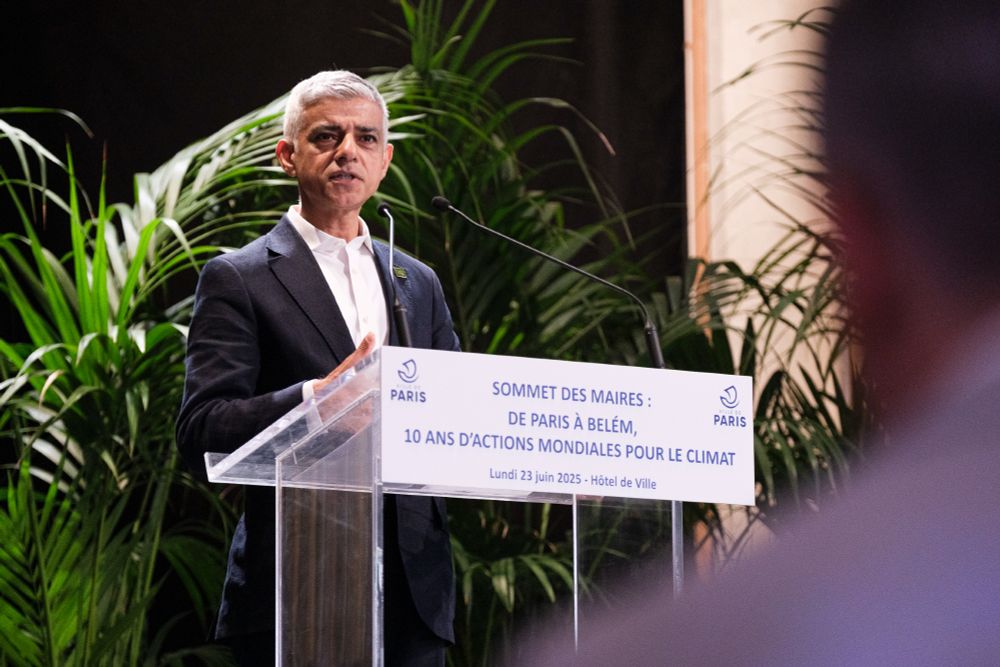 The Mayor of London speaks at a podium at the Paris Summit of Mayors, with greenery in the background.