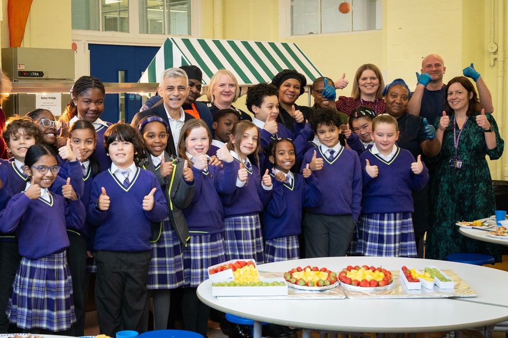 
The Mayor of London and a large group of students in purple uniforms posing with thumbs up around a table with platters of fruit spelling out "100M"

