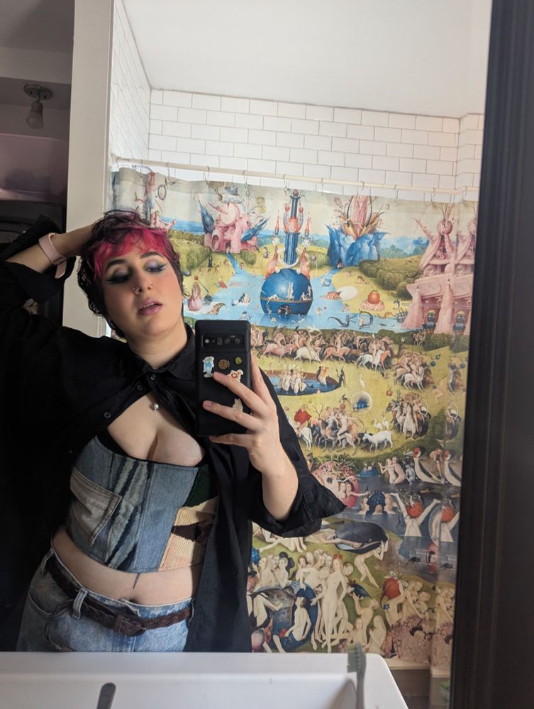 Syd, a white non-binary person with dark brown and pink hair stares into the camera. They are wearing green, blue and silvery makeup with lots of highlighter. They sport a black button up and jean corset. They're also wearing a little pearl necklace with star shaped nose piercings. They stare into a bathroom mirror with their phone as they pose. Their shower curtain is garden of earthly delights by Hieronymus Bosch.