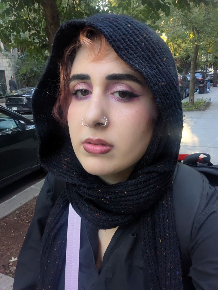 Syd stares tiredly at the camera. They're wearing a black scarf/shawl wrapped around their head. They are sporting pink eyeshadow and double star piercings on their nose.