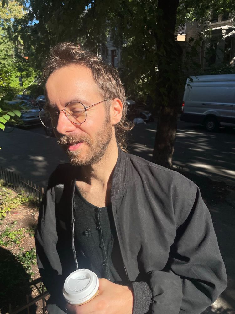 Elio, who is currently experiencing their birthday, is dressed in all black and clutching a coffee cup. They're wearing lil glasses and closing their eyes as the sun hits them.