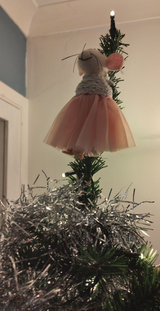 Mouse in a pink tutu on top of a Christmas tree. She's held in place by her pipe cleaner arms around the trunk, so it looks a bit like she's tied to the tree.
