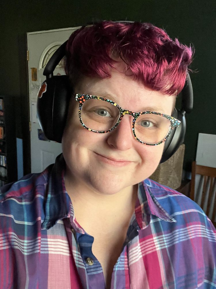 Photo of k morr transmasc person with glasses with floral frames dark pink short hair wearing headphones and a blue and dark pink plaid shirt. They are smiling in a genuine but bit dorky way.