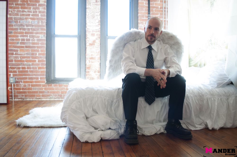A bald man in a white shirt, black tie, and black pants sitting on a bed in front of backlit windows wearing angel wings. 