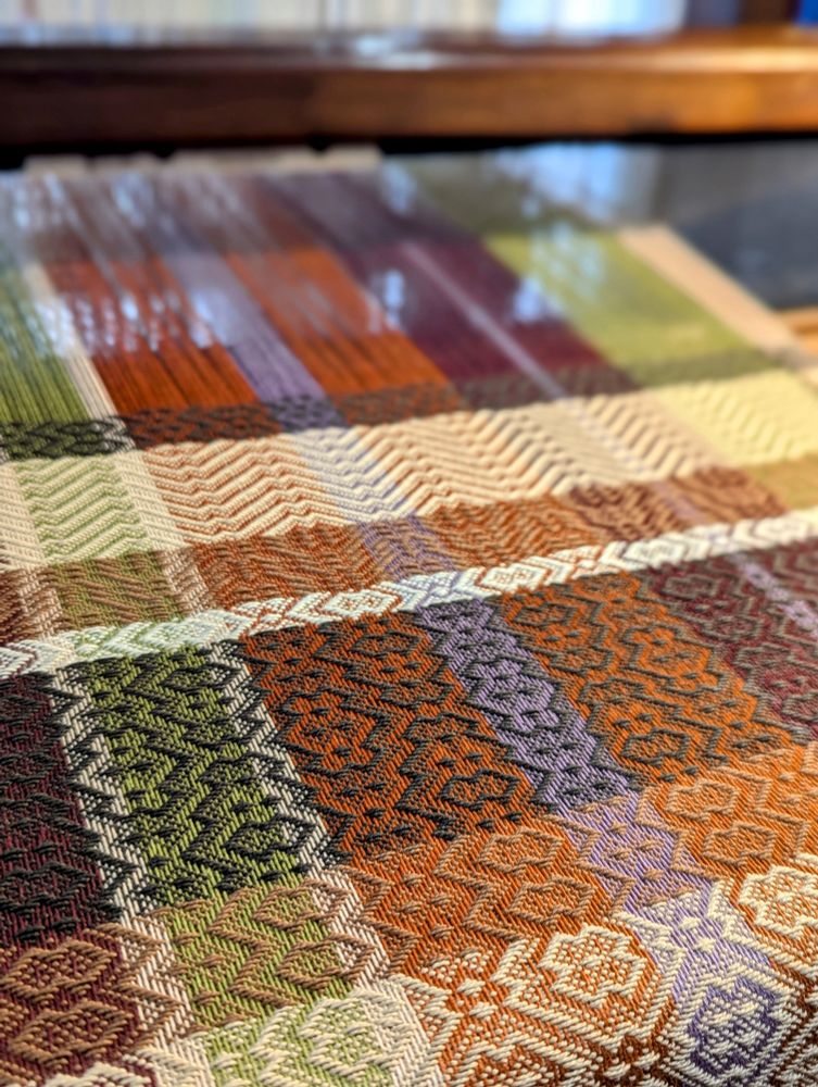 Striped warp in shades of burgundy, orange, green, lavender, cream on a wooden countermarche floor loom. Weft is woven with stripes of beige, brown and cream. Pattern is 8 shaft M's and W's twill, woven in 8/2 cotton for tea towels.