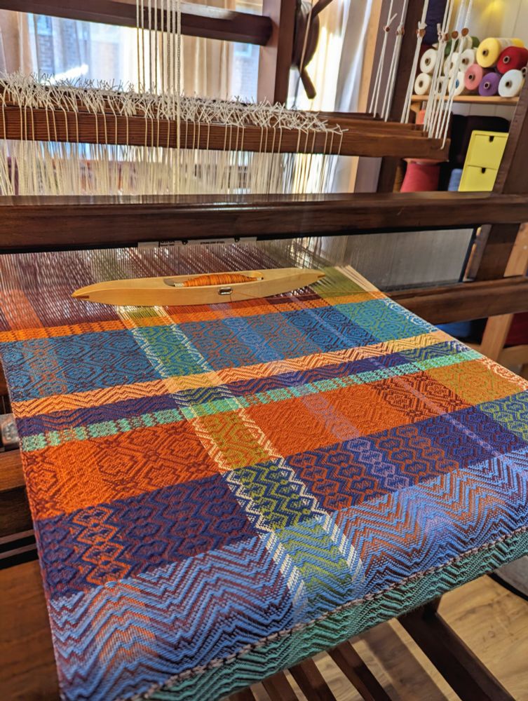 Colourful striped warp being woven on a wooden 8 shaft loom into tea towels with intersecting orange and blue horizontal and vertical stripes. 