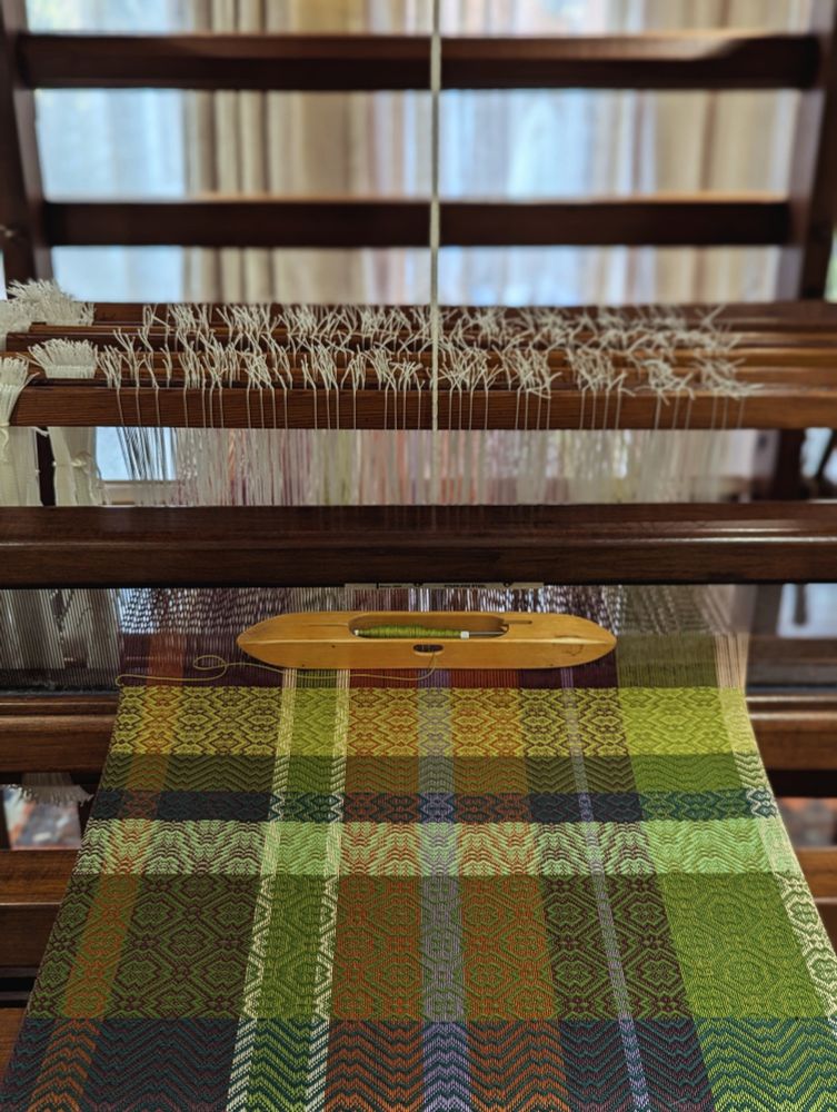 Tea towel being woven on an old wooden floor loom. Cloth is being woven in intersecting horizontal and vertical bands of green, orange, white, purple and maroon. A wooden boat shuttle is currently filled with a bobbin of pale green thread. 