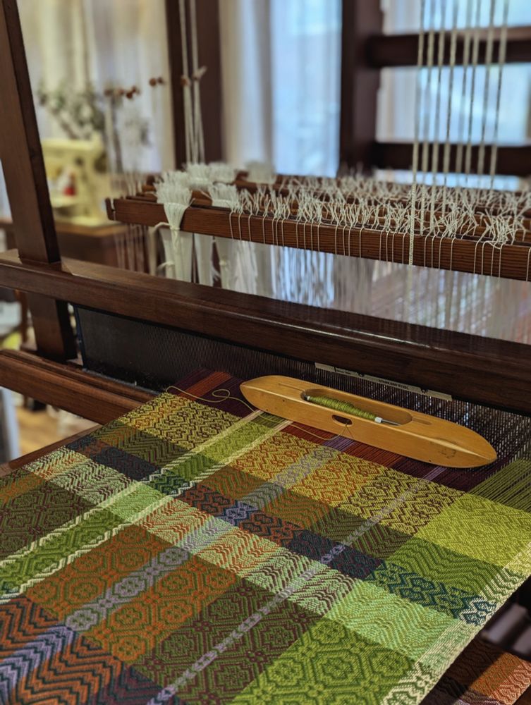 Tea towel being woven on an old wooden floor loom. Cloth is being woven in intersecting horizontal and vertical bands of green, orange, white, purple and maroon. A wooden boat shuttle is currently filled with a bobbin of pale green thread. In the background you can see windows and an old sewing machine. 