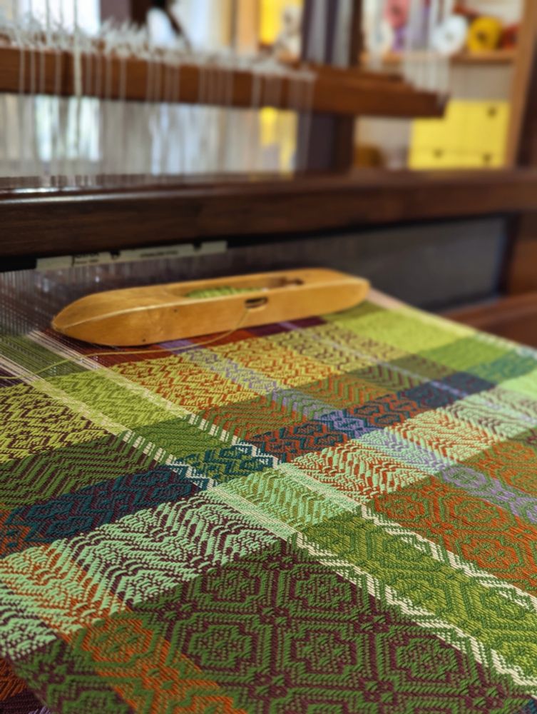 Tea towel being woven on an old wooden floor loom. Cloth is being woven in intersecting horizontal and vertical bands of green, orange, white, purple and maroon. A wooden boat shuttle is currently filled with a bobbin of pale green thread. 