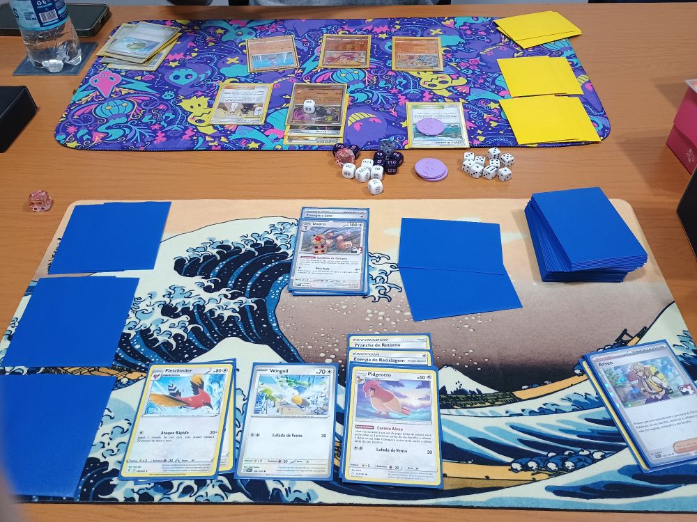 Foto de uma visão em primeira pessoa de uma partida de Pokémon TCG. Virado para o observador da foto um baralho com vários pokémons passarinhos, do outro lado da mesa um baralho com pokémons lutadores.