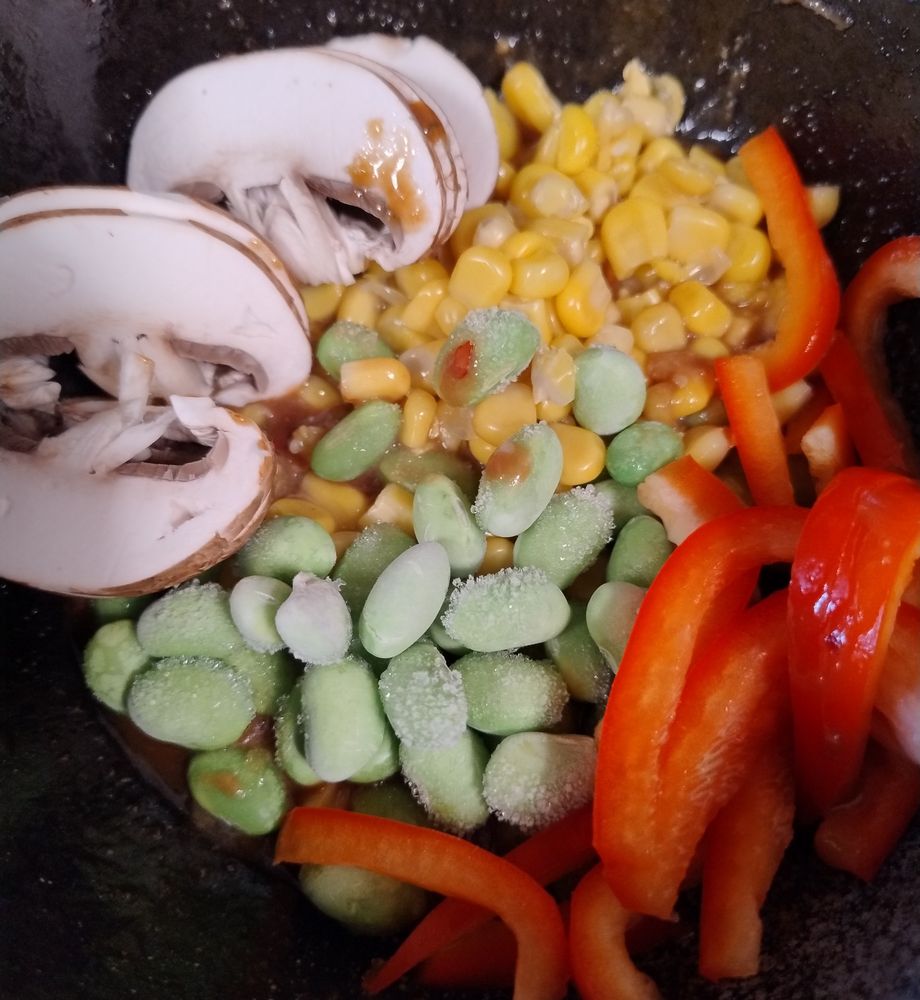 In einer Schüssel Mais, gefrorene Edamame, Paprikastreifen, Champignonscheiben, darunter ist bräunliche Sauce zu ahnen.