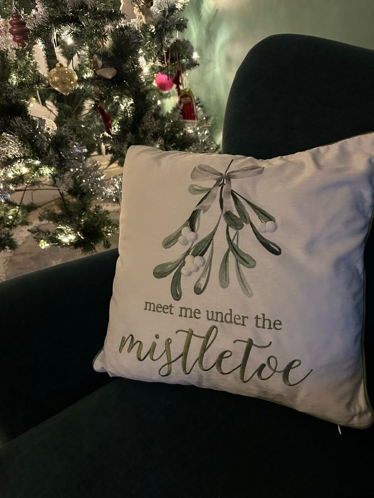 A white and green cushion with meet me under the mistletoe in it . Green mistletoe with white bobble berries 