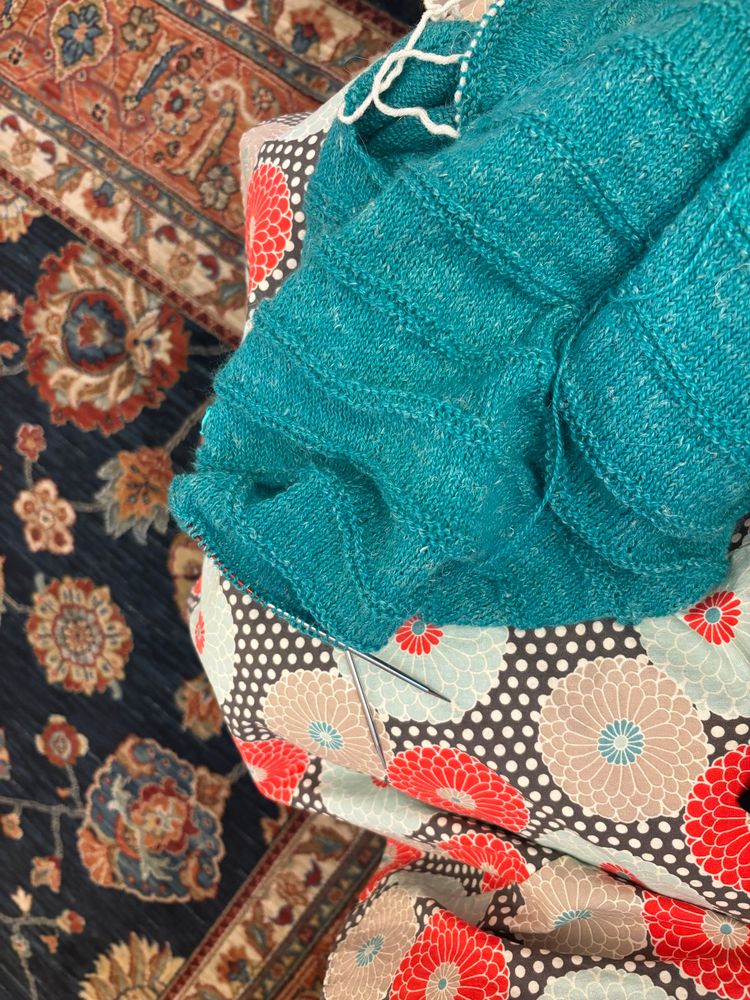 Turquoise sweatee being knit. It rests on a person’s lap who is wearing a skirt with flowers and polka dots. The rug in the background also has flowers on it. 