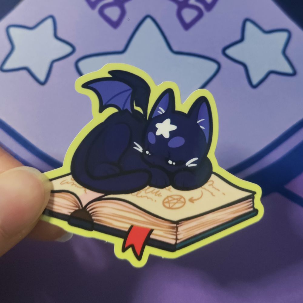 Photo du sticker d'octobre: il représente un chat noir avec des ailes de chauve-souris et une étoile blanche sur le front. Il est en boule endormi sur un grimoire 