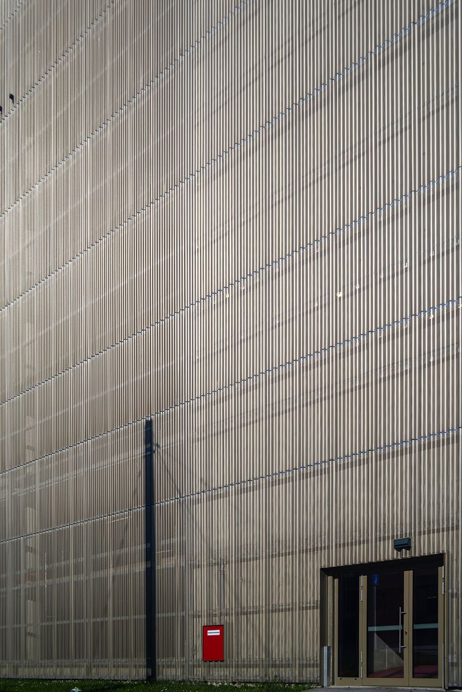 A minimalist architectural photograph of a tall, modern warehouse wall with prominent vertical corrugated metal siding. A sharp diagonal shadow cuts across the surface from a vertical pole on the left. Towards the bottom right, a modern glass double door entrance with reflections is visible, and next to it, a small red box stands out.