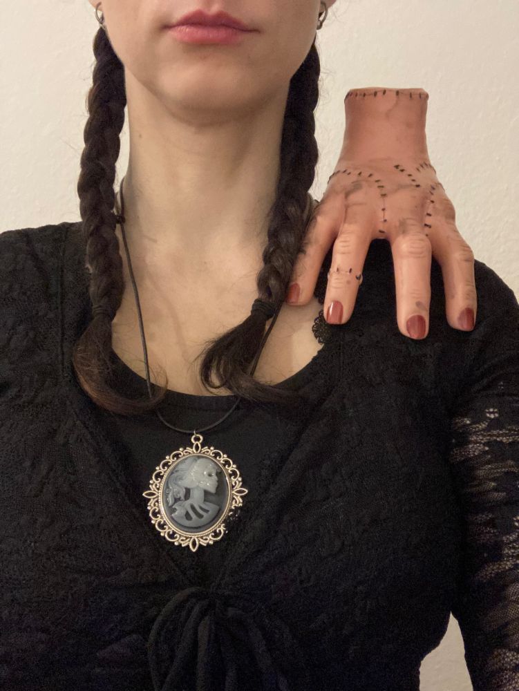 Zu sehen ist eine Person von ab unter der Nase bis zum Großteil des Brustkorbs. Die Person hat zwei schwarze geflochtene Zöpfe, ist schwarz gekleidet mit einer Kette, auf deren Anhänger eine Skelett-Person zum Teil zu sehen ist. Auf der Schulter sitzt eine Hand, die mehrere Nähte besitzt.