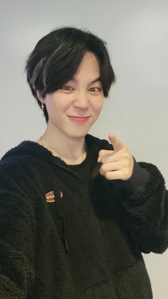 Jimin in a black hoodie with highlights in his hair posing for a selfie with his nose scrunched and a finger pointing towards the camera