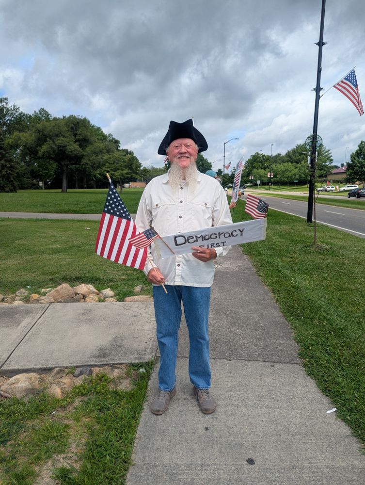 A smiling man standing on a sidewalk and wearing a revolutionary tricorn hat carrying an American flag and a sign that says Democracy First