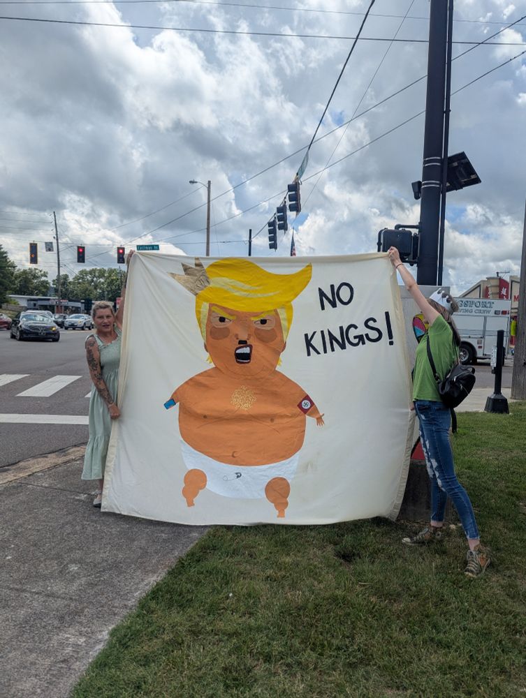 Very large sign with an image of a baby Trump in a diaper that says No Kings! Held by two protesters on a street corner