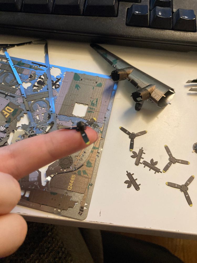 A single finger outstretched with a miniature plane propellor balanced on the end. It is painted black and yellow and made of thin metal. In the background, a miniature steel model kit of a B-17 Flying Fortress is strewn across a white desk, one wing partially constructed and three other propellers waiting to be assembled. 