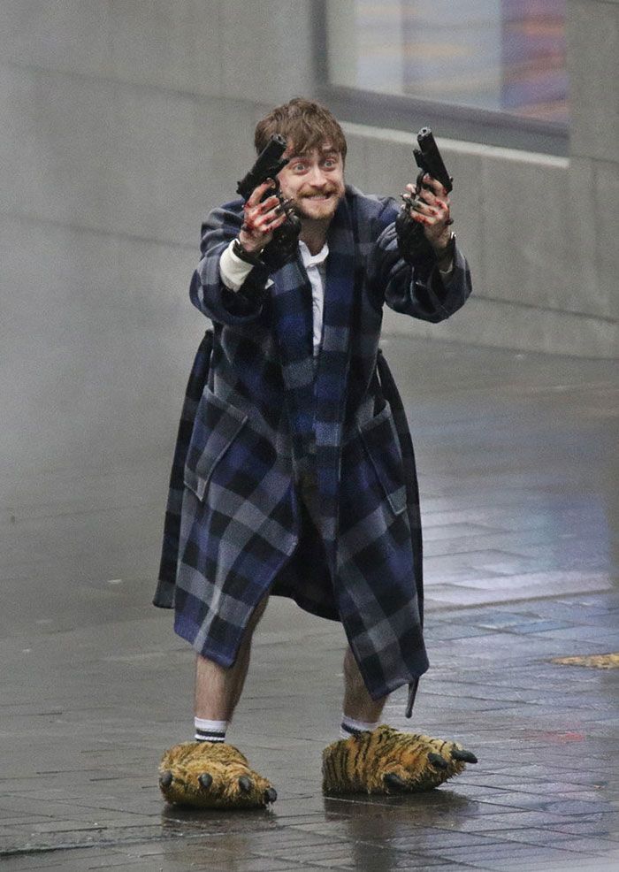 A photo of actor Daniel Radcliffe holding two handguns, one in each hand, while on a wet city street. He is smiling mischievously and looking at the camera. He's dressed in a plaid bathrobe, a white T-shirt, and what appear to be oversized tiger-claw slippers. His white socks are pulled up to his calves, and his legs are bare. There is blood on his right hand and the cuffs of his shirt. In the background, a large building with a concrete or stone facade is visible, though it is slightly out of focus.