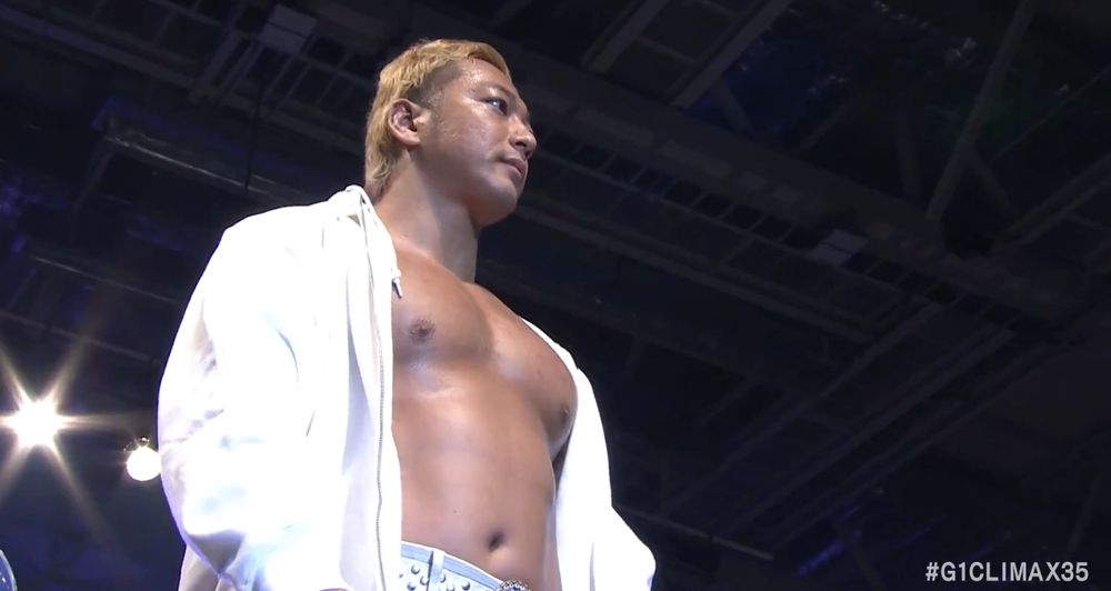 A screenshot of Shota Umino standing on the turnbuckle, looking onward breastily.