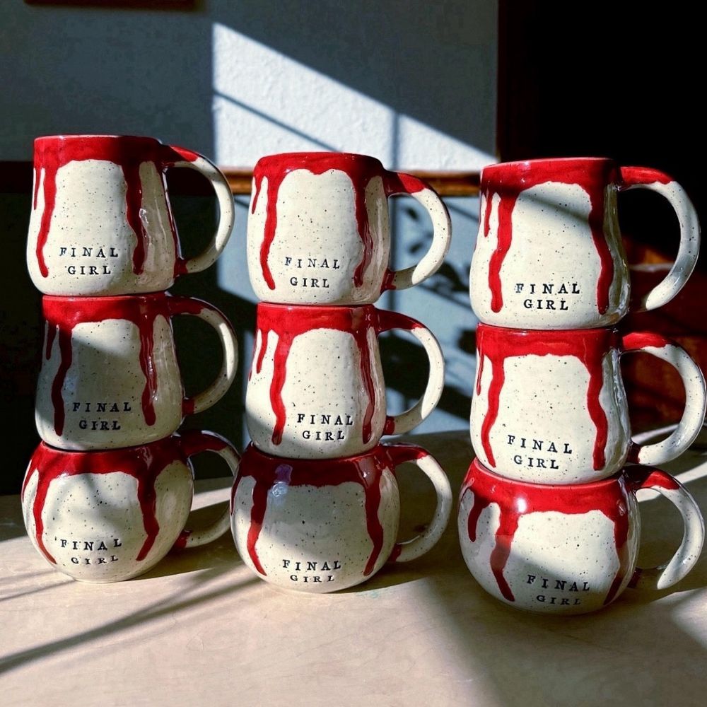 Nine handmade pottery mugs that read FINAL GIRL stacked 3 by 3 on a table. The mugs are made from speckled white clay and glazed blood red inside with blood red dripping down the outsides. 