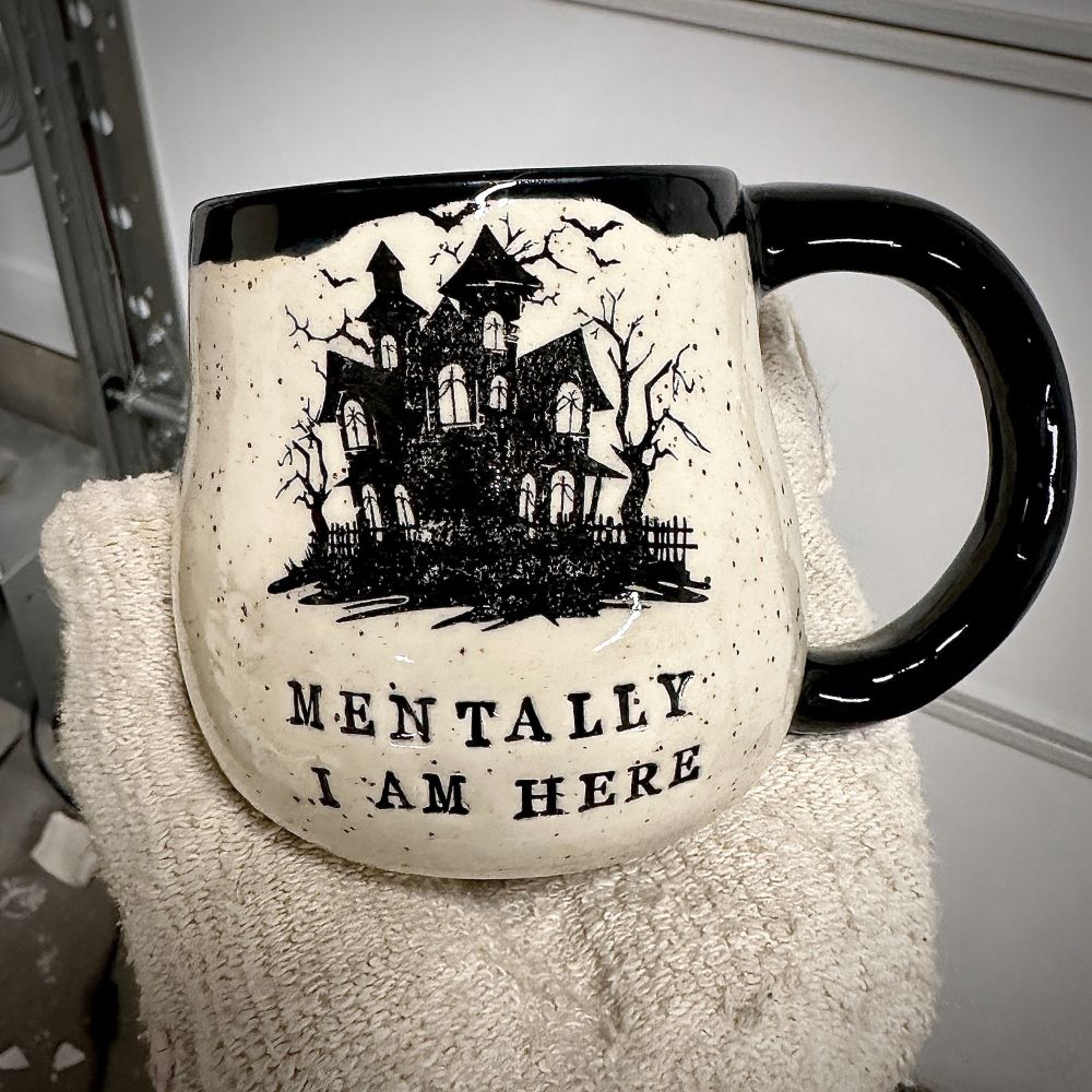 A mitted hand holding up a white speckled clay mug that reads “MENTALLY I AM HERE” in black letters with a black image of a haunted house. The interior, handle, and rim are glazed glossy black. 