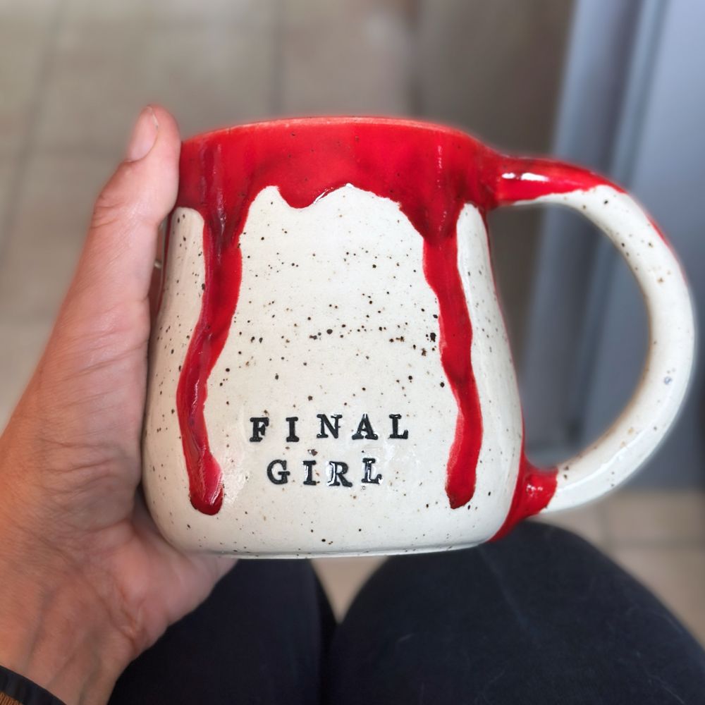 A hand holding up a white speckled clay handmade pottery mug that reads FINAL GIRL. The mug is glazed blood red on the inside and dripping down the outsides. 