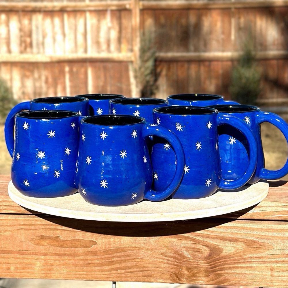 Eight royal blue handmade pottery mugs glazed black on the inside with small white stars carved on the outside. The mugs are resting on a wooden bat on top of a backyard fence. 