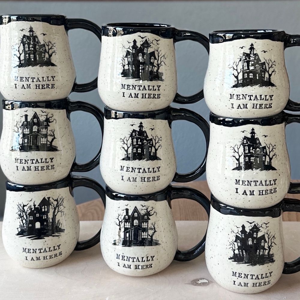 Nine speckled white clay handmade pottery mugs stacked 3 by 3 that have an image of a haunted house and read MENTALLY I AM HERE. The mugs are glazed clear on the outside and glossy black on the inside, handle, and rim. 