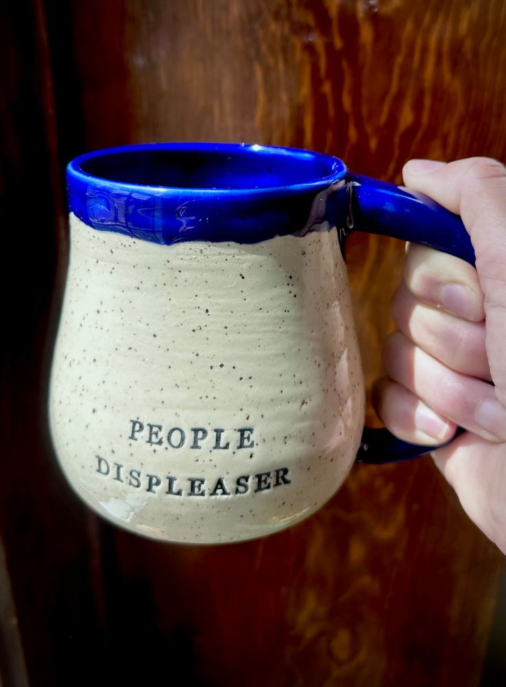 A white speckled clay handmade pottery mug that reads PEOPLE DISPLEASER. The mug is glazed cobalt blue on the handle, rim, and interior. 