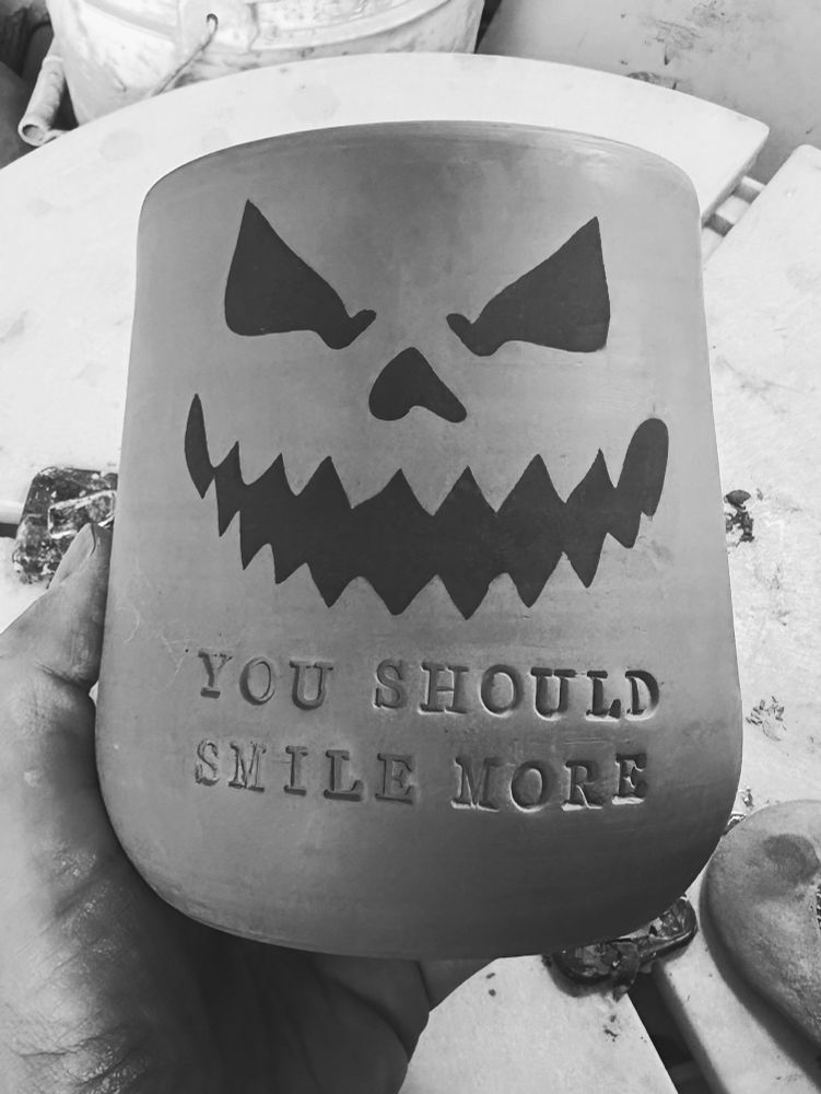 A handmade pottery mug in my hand that reads YOU SHOULD SMILE MORE and has a black jack-o’-lantern face. The photo is black and white. 