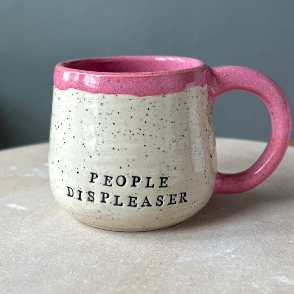 A white speckled handmade pottery mug that reads PEOPLE DISPLEASER. It’s glazed hot pink on the inside, handle, and rim. 