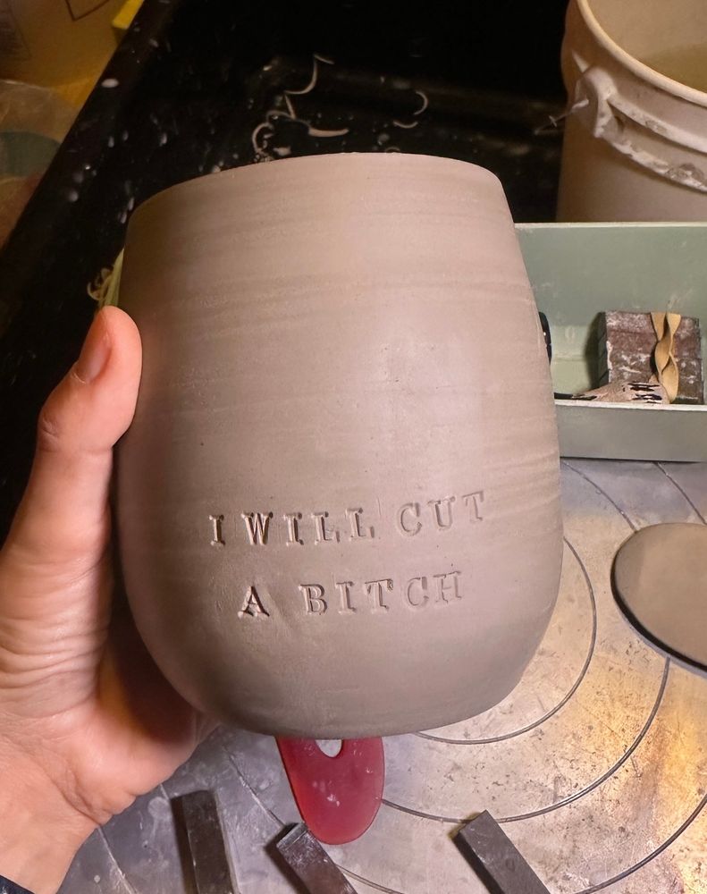 A leatherhard white clay pottery mug that reads “I WILL CUT A BITCH” held in my hand over a pottery wheel 