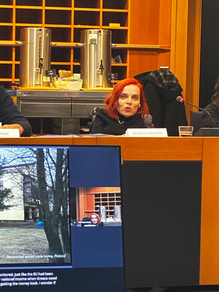 A woman in a wheelchair with red hair wearing a black blazer speaks at a microphone during a European Parliament hearing. She sits at an orange desk with a nameplate reading "ANTONIA TRIKALIOTI" in front of her. Behind her is a beverage station with stainless steel dispensers and orange-paneled shelving. On a monitor below, a video presentation shows a renovated social care home in Poland, with caption text visible discussing EU monitoring and national income.