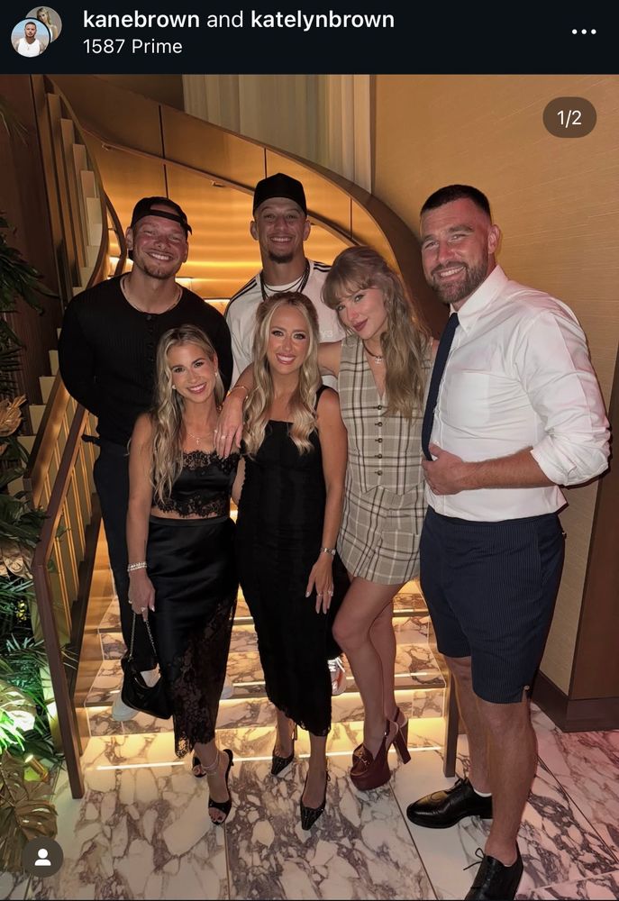 Photo of a group of people standing in front of/on bottom steps of spiraling staircase. Taylor Swift is featured in the front row of people, wearing plaid two piece vest and skirt set with red Louis Vuitton horsebit platforms