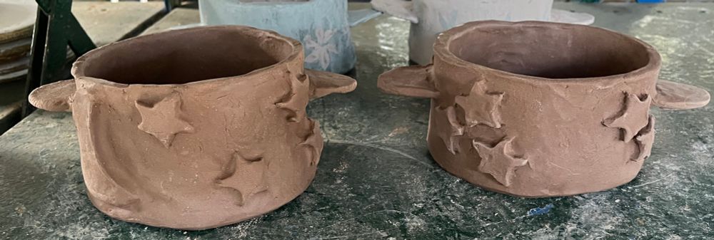 2 Ceramic small round pots made of brown clay. Pot 1is decorated with stars and a crescent moon. Pot 2 is decorated with stars.The pots have not been fired or glazed.