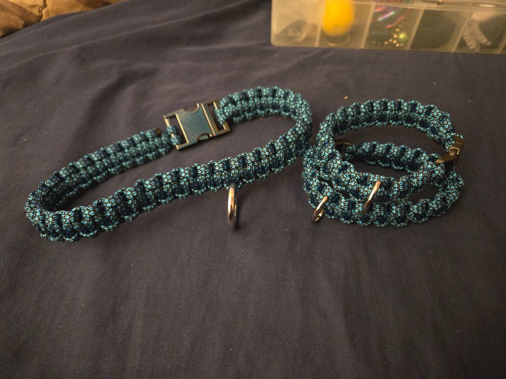 A collar and cuff set sitting on a bed. The collar is on the left and the cuffs are stacked on the right. The paracord is a blue and black weave colour. 