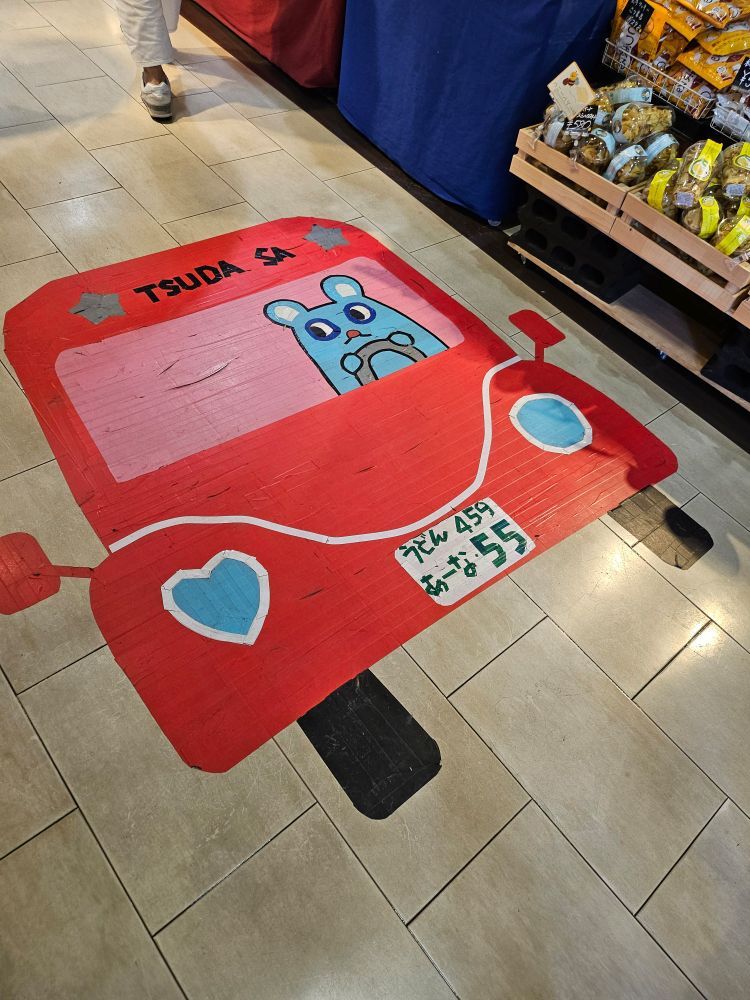 [写真]床にカラフルなテープで描かれたイラスト。赤い車に乗った水色のネズミのような生き物。