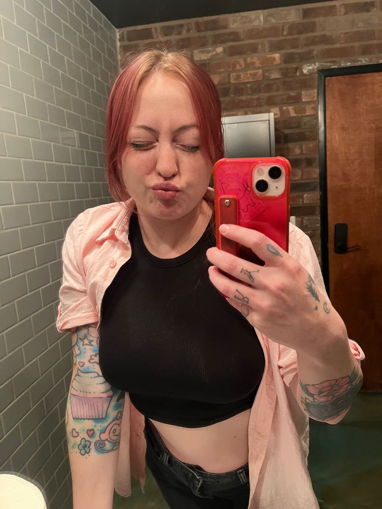 A pale woman with a hot pink case is wearing a black crop top, a pink button up over it, and black jeans is leaning into the mirror with lips puckered and eyes closed.