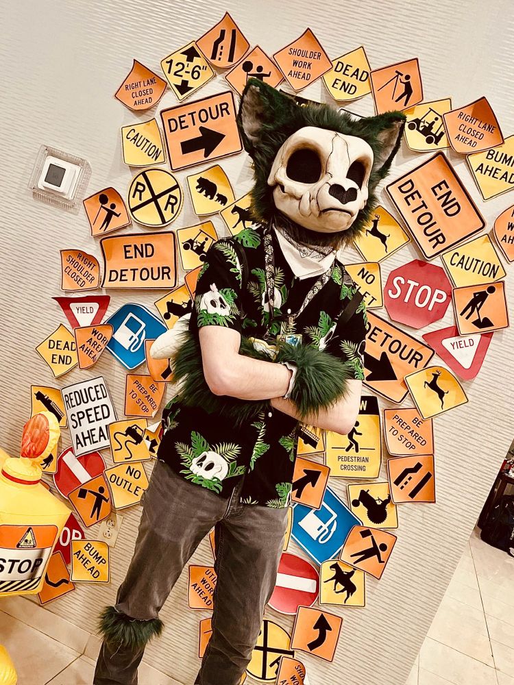 green and bone colored skullbeast fursuiter crossing arms in front of a collage of road signs