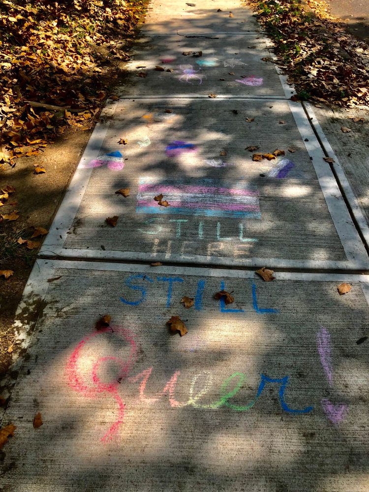 A chalk drawing of a trans flag with the words “still here, still queer” 