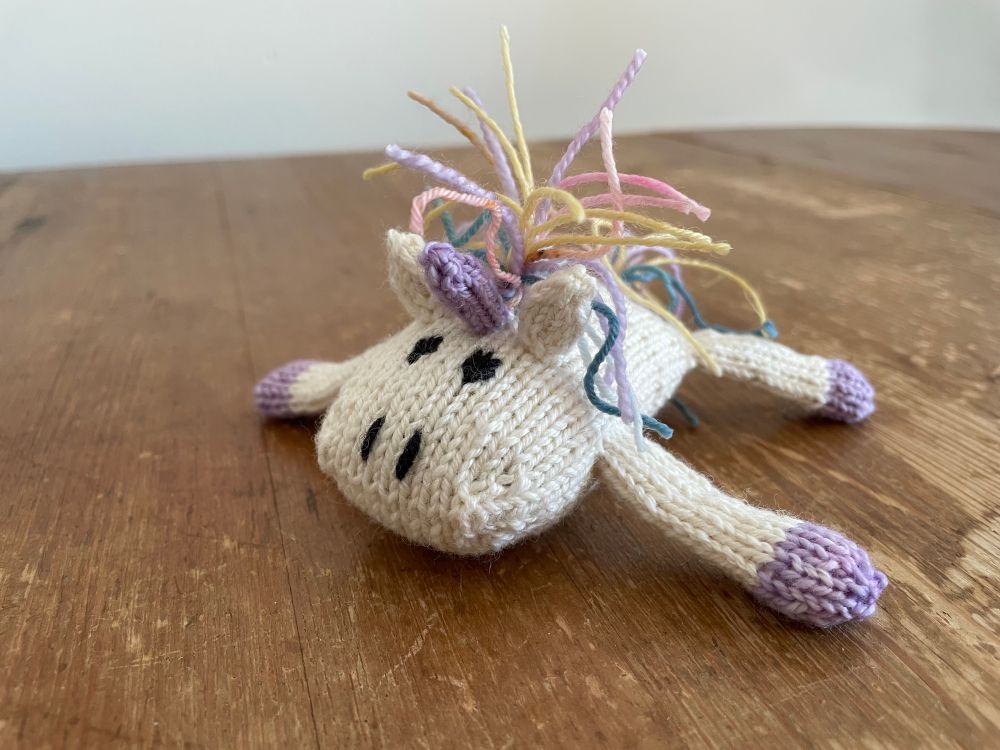 a hand-knit unicorn, with lilac horn and hooves and a delightful pastel-shaded mane. his body is very flat and his legs are splayed out from his body.